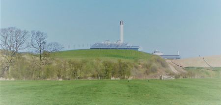 Green fields and rolling hills stand in front of grey-cladded buildings with a tall chimney.